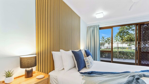 A modern bedroom with a large bed, wooden headboard, bedside table with a lamp, and a view of the outdoors through a sliding door.