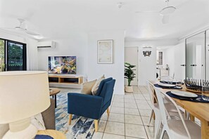 A modern living room with a blue sofa, a TV, and a dining area with a wooden table and chairs. - Oceanfront 3 Bedroom Apartment with Pool in Cairns (Cairns North)