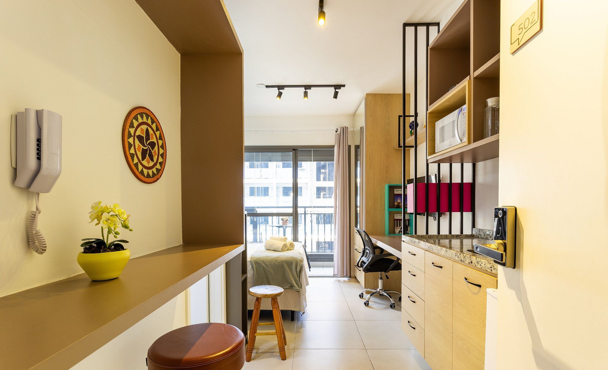 Quarto de hotel compacto com cozinha compacta, cama com vista, um banquinho pequeno e uma planta em vaso.