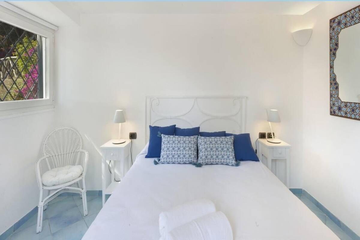 Un dormitorio blanco con una cama, dos mesitas de noche con lámparas, una silla blanca y un ventanal con vistas decorativas.