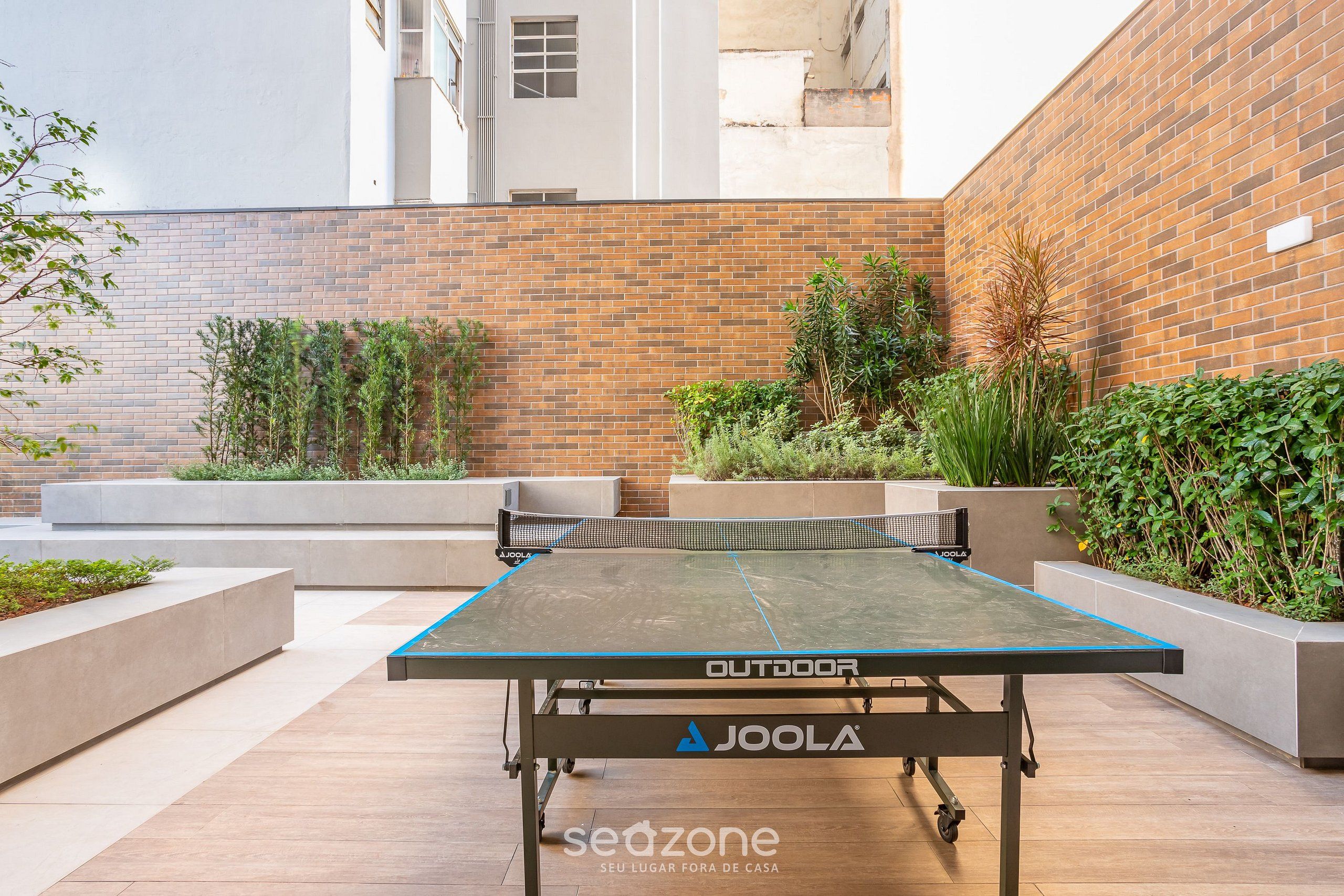 Un tavolo da ping pong all'aperto con il marchio 'Joola' e un logo 'seazone', circondato da fioriere e un muro di mattoni.