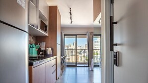 A modern kitchen with a refrigerator, microwave, and wooden cabinets. A dining area with chairs and a table is visible. The room has a large sliding glass door leading to a balcony with a city view. - ISC - Well-located Studios in Vila Buarque, Sao Paulo (São Paulo)