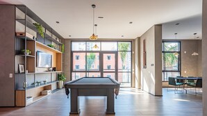 A modern living room with a pool table, built-in shelving with plants, large windows, and a dining area with green chairs. - ISC - Well-located Studios in Vila Buarque, Sao Paulo (São Paulo)