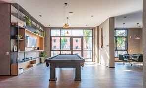 Ein modernes Wohnzimmer mit Billardtisch, integrierten Regalen mit Pflanzen, großen Fenstern und einem Essbereich mit grünen Stühlen.