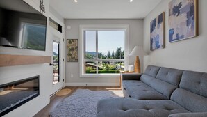 A modern living room with a grey sofa, a fireplace, a large window with a view of greenery and mountains, and abstract art on the walls. - Harrison Hot Springs Lakeside. One Bedroom Apartment, Balcony and Mountain View. (Harrison Hot Springs)