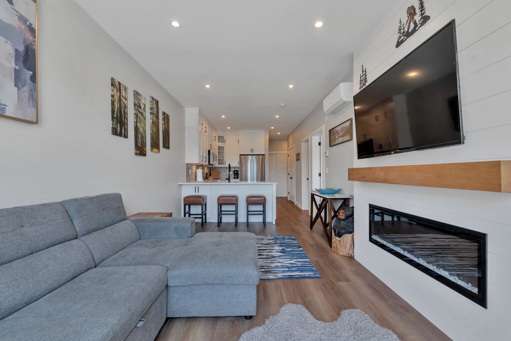 A modern living room with a grey sectional sofa, a fireplace, and a kitchen in the background.