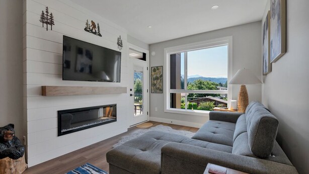 A modern living room with a gray sofa, a flat-screen TV mounted on a white wall, a fireplace, and a large window offering a view of greenery and mountains. - Harrison Hot Springs Lakeside. One Bedroom Apartment, Balcony and Mountain View. (Harrison Hot Springs)
