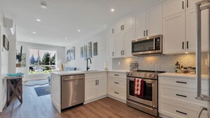 A modern kitchen with white cabinets, stainless steel appliances, and a large window offering a view of trees. - Harrison Hot Springs Lakeside. One Bedroom Apartment, Balcony and Mountain View. (Harrison Hot Springs)