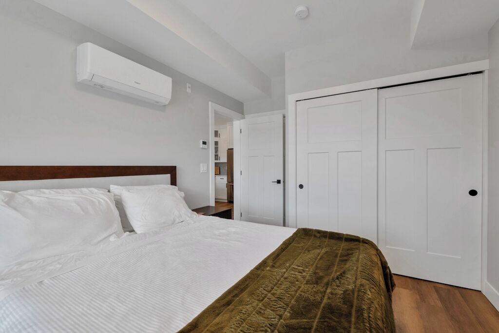 A bedroom with a bed, white walls, a wooden headboard, a brown throw blanket, and white doors.