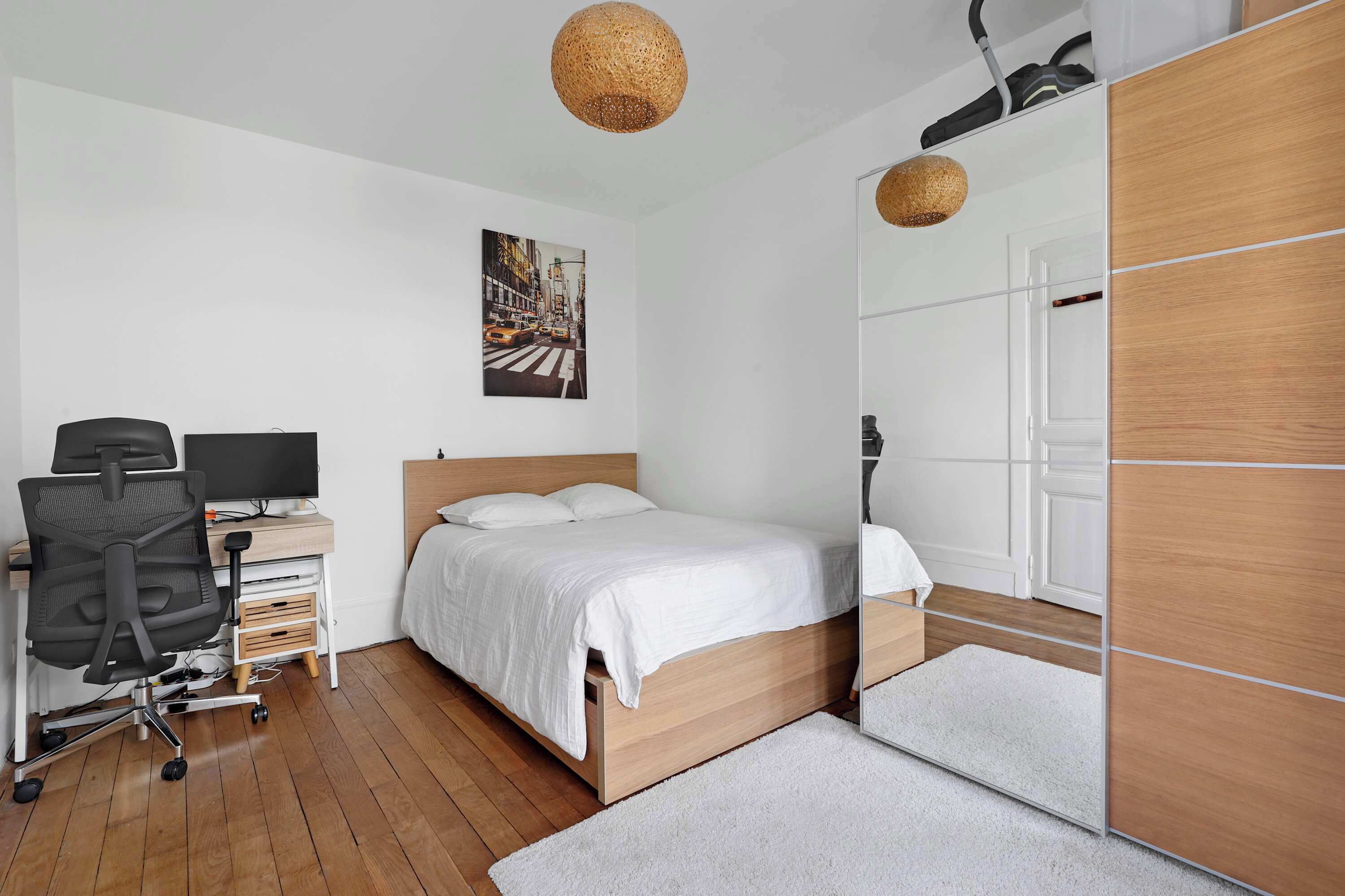 Une chambre moderne avec un lit en bois, un bureau avec une chaise, un grand miroir et une œuvre d’art représentant un paysage urbain accrochée au mur.