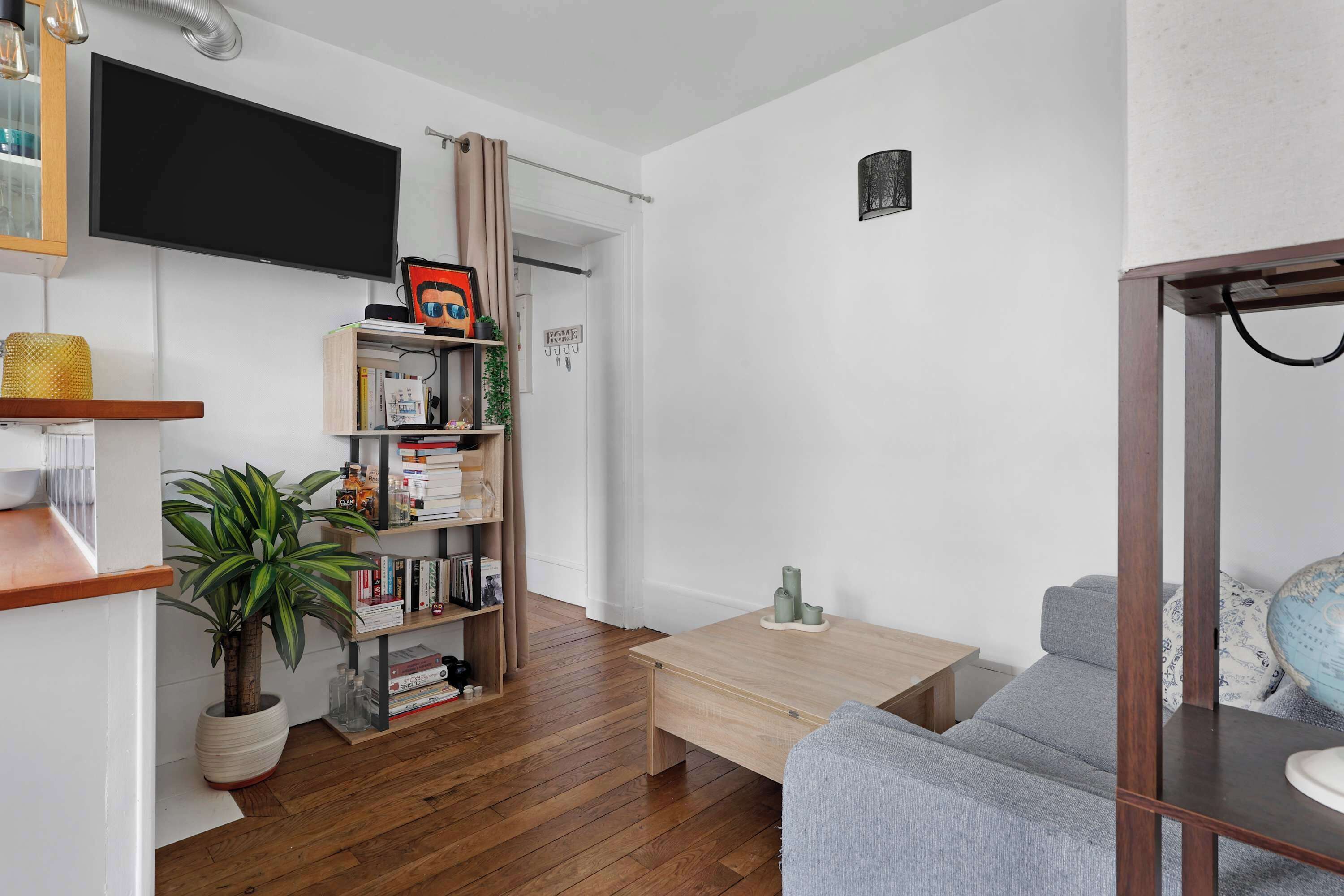 Un salon avec un plancher en bois, un canapé gris, un téléviseur fixé au mur, une bibliothèque et une plante en pot.