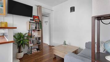 Un salon avec un plancher en bois, un canapé gris, un téléviseur fixé au mur, une bibliothèque et une plante en pot.