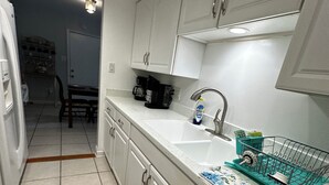 A compact kitchen with white cabinets, a built-in dishwasher, and a countertop with a dish rack. - The Josephine  8 Beds (Fort Walton Beach)