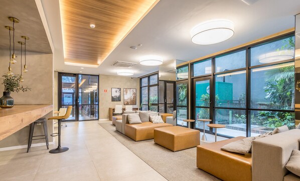 A modern lounge area with a wooden reception desk, comfortable seating, large windows, and a view of the outdoors. - Modern Comfort 450m From the Subway Ism0806 (São Paulo)