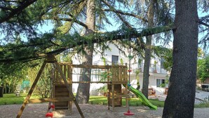 A playground with a wooden swing set and slide, surrounded by trees and a building in the background.