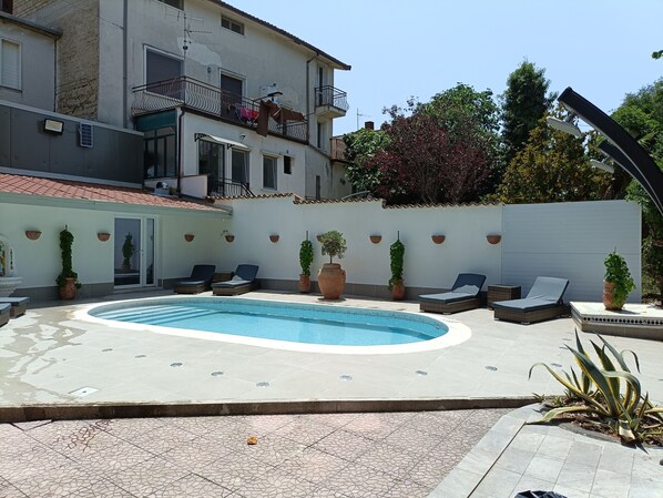 Une piscine entourée de chaises longues et de plantes en pot, avec un bâtiment en arrière-plan.