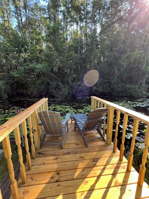 Una terraza de madera con sillas Adirondack y una mesa, rodeada por un bosque denso y un estanque con nenúfares.