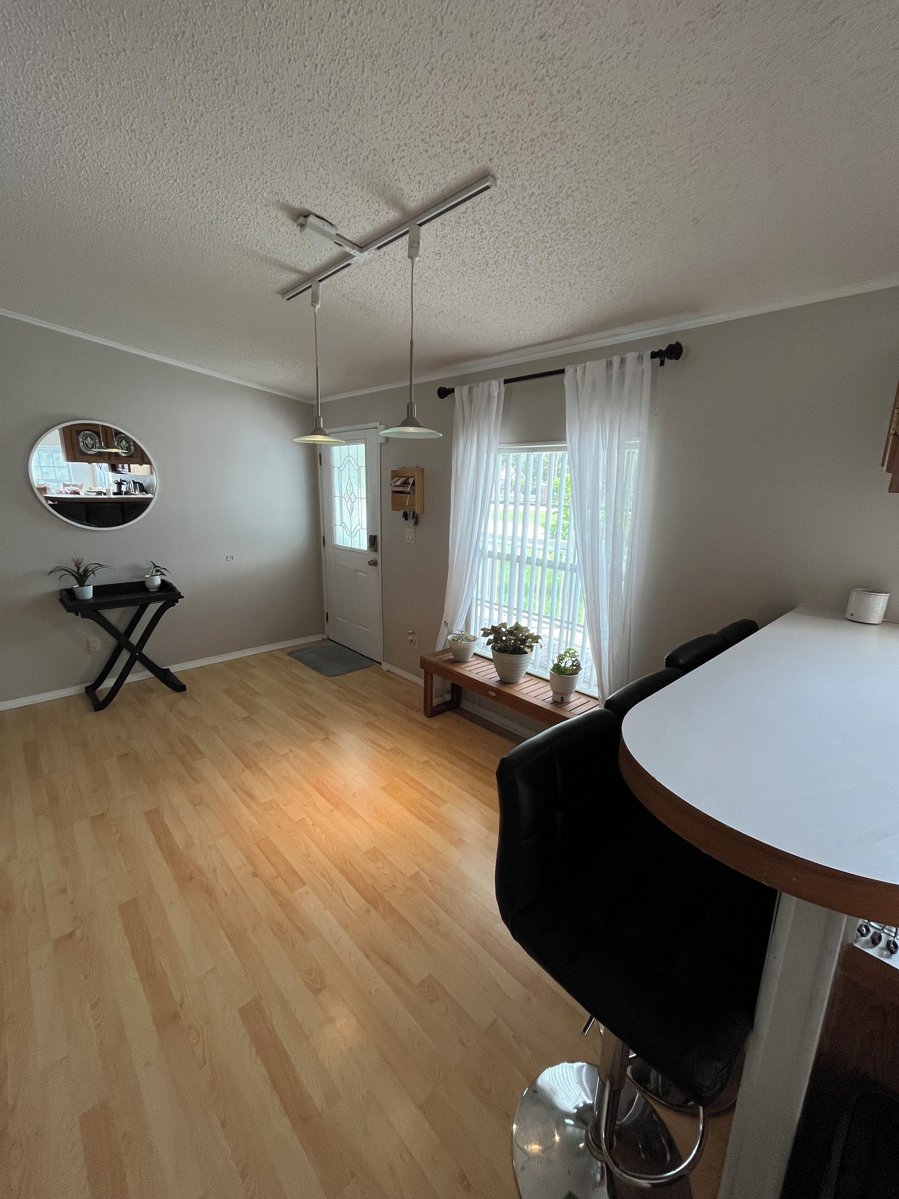 Una habitación con piso de madera, un espejo redondo en la pared, una mesita con plantas en macetas, una mesa blanca y una silla negra.