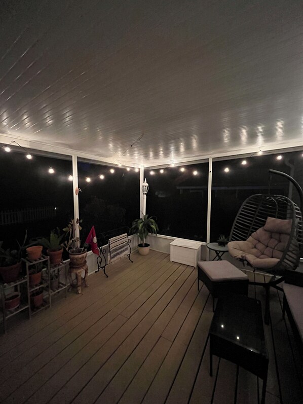 Un espacio al aire libre cubierto con cubierta de madera, macetas, un banco, una silla y luces colgantes.