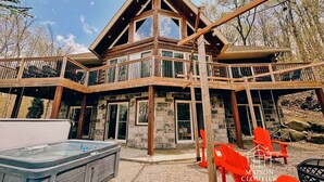 A two-story log cabin with a balcony, stone foundation, and a hot tub in the foreground.