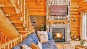 A log cabin interior with a stone fireplace, a blue sofa, and a wooden staircase.