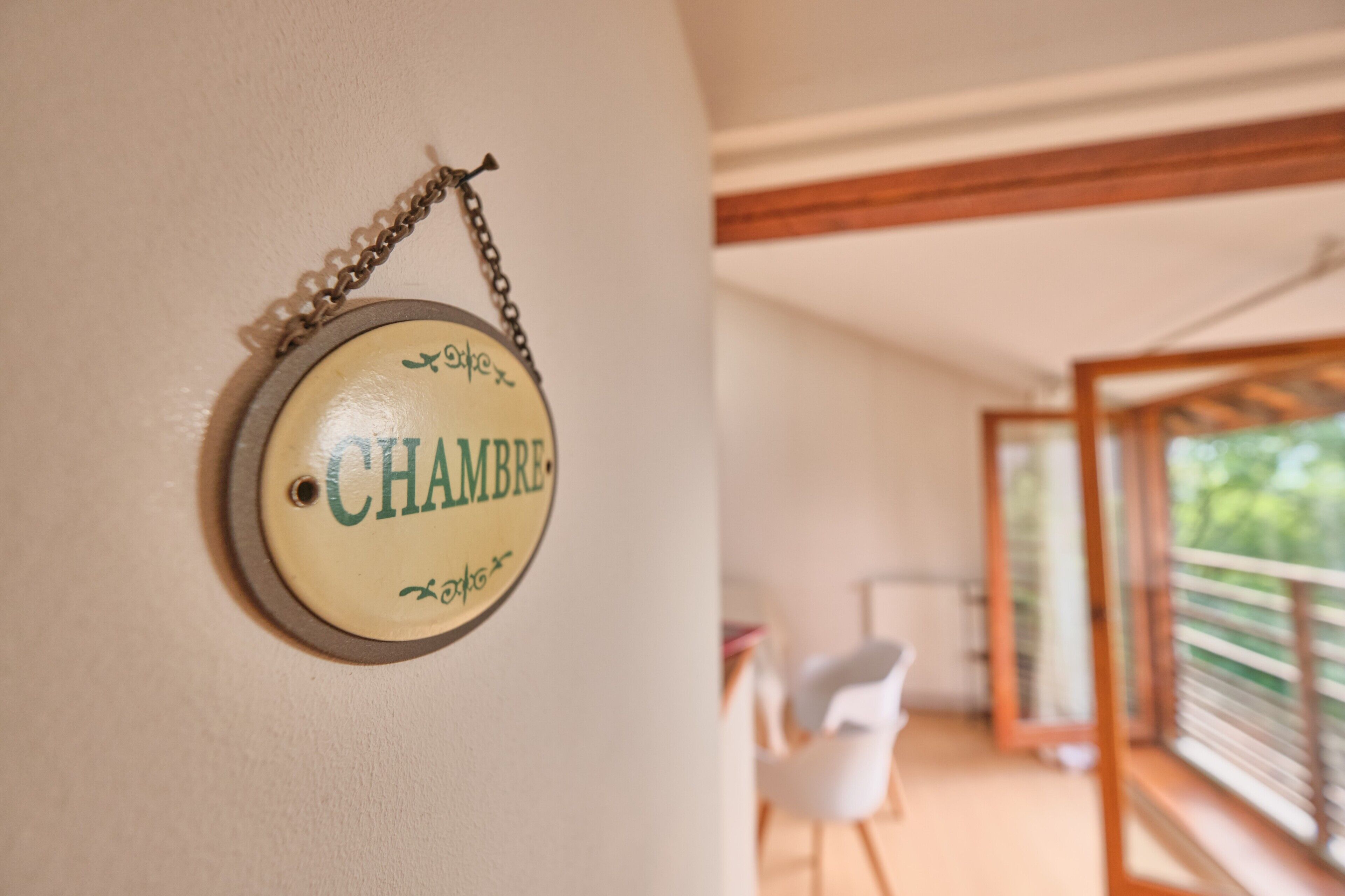 Ein rundes Schild mit dem Wort 'CHAMBRE' an einer Wand, ein weißer Stuhl und der Blick auf einen Balkon mit Geländer.