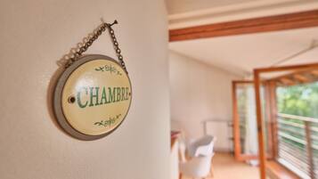 Ein rundes Schild mit dem Wort 'CHAMBRE' an einer Wand, ein weißer Stuhl und der Blick auf einen Balkon mit Geländer.