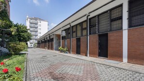 A paved walkway with potted plants and red flowers, a building with a brick facade and large windows, and a taller building in the background. - Room 'Tripla' with Wi-Fi and Air Conditioning (Milano)