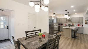 Una cocina moderna con una isla central, iluminación colgante y una zona de comedor con una mesa y sillas de madera oscura.
