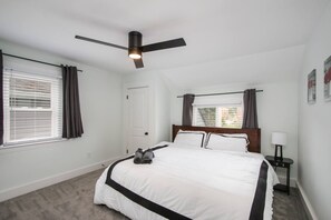 A bedroom with a bed, a ceiling fan, a window with curtains, a door, and a nightstand with a lamp.