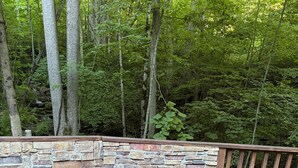 A stone fire pit with a fire, surrounded by a wooden deck and a stone wall, with chairs and a wooden railing. - Creekside Retreat-Close to Sylva, Cherokee, and Dillsboro (Whittier)