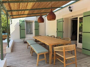 Een terras buiten met een houten tafel en banken, groene luiken en een geweven dak.