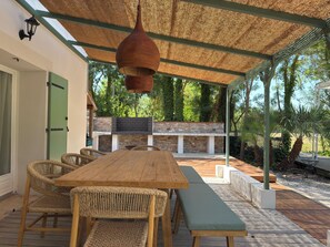 Een terras buiten met een houten tafel en rieten stoelen, een hanglamp en een stenen openhaard.