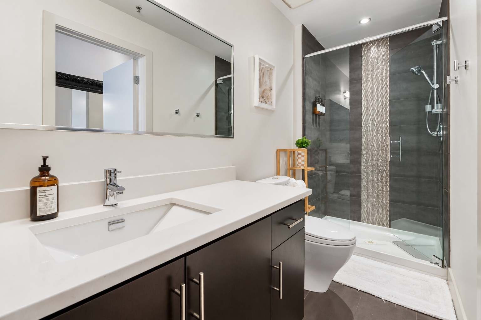 A modern bathroom with a large mirror, a white sink, dark cabinets, a toilet, and a glass-enclosed shower area.
