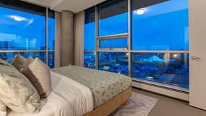 A bedroom with a large bed, multiple pillows, and a view of a cityscape through large windows. - 2-Storey PH Pano Views+2 Terraces|Parking|Soaker Tub (Calgary)