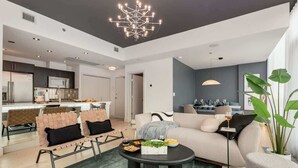 A modern living room with a sofa, coffee table, and chairs. There is a kitchen in the background with a microwave and cabinets. - 2-Storey PH Pano Views+2 Terraces|Parking|Soaker Tub (Calgary)