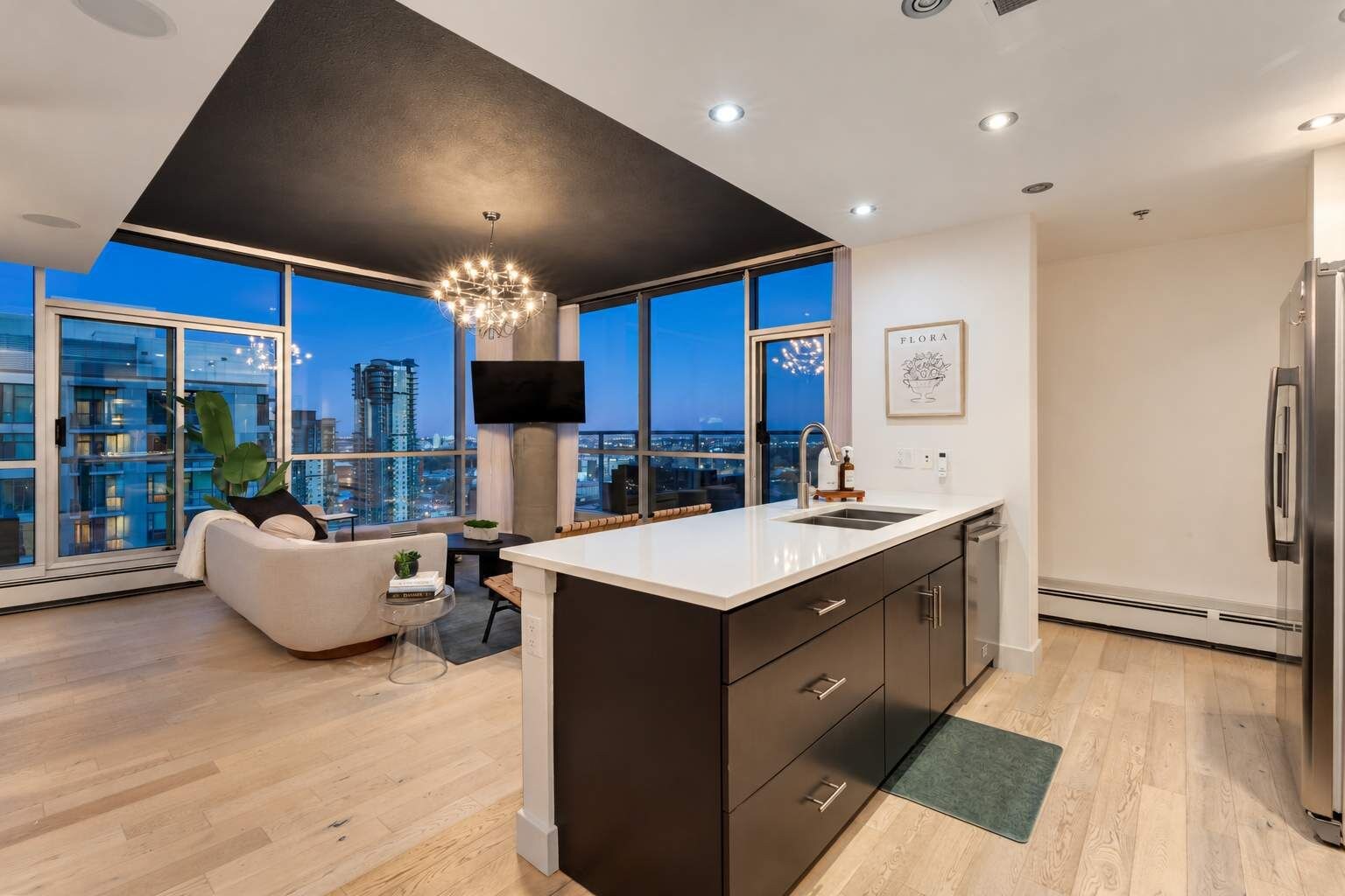 A modern kitchen with a large island, a dining area with a sofa, and a city view through large windows.