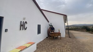 A building with a sign 'HC' on the wall, a paved courtyard, and a view of distant hills. - Holiday Home 'Herdade Da Carapuça' with Mountain View, Private Pool and Wi-Fi (Portalegre)