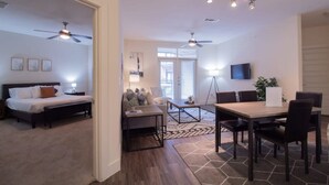 A modern living room with a sofa, coffee table, and dining table. There are ceiling fans, a TV, and a window with blinds. - Revive Tanglewood Stunning 2BR in Uptown Houston (Houston)
