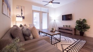A modern living room with a sofa, coffee table, and a patterned rug. There is a ceiling fan, a TV mounted on the wall, and a potted plant. - Revive Tanglewood Stunning 2BR in Uptown Houston (Houston)