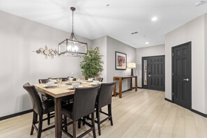 Un coin repas avec une table en bois, des chaises sombres, un luminaire suspendu, une plante en pot et une porte en arrière-plan.