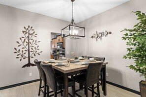 A dining area with a wooden table, black chairs, a hanging light fixture, and wall decorations including a tree and a bird. - Waters Edge Retreat – For 6! (Branson)
