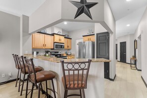 Une cuisine moderne avec des meubles en bois, un îlot central doté de tabourets de bar, un réfrigérateur en acier inoxydable et une décoration en forme d’étoile noire au plafond.