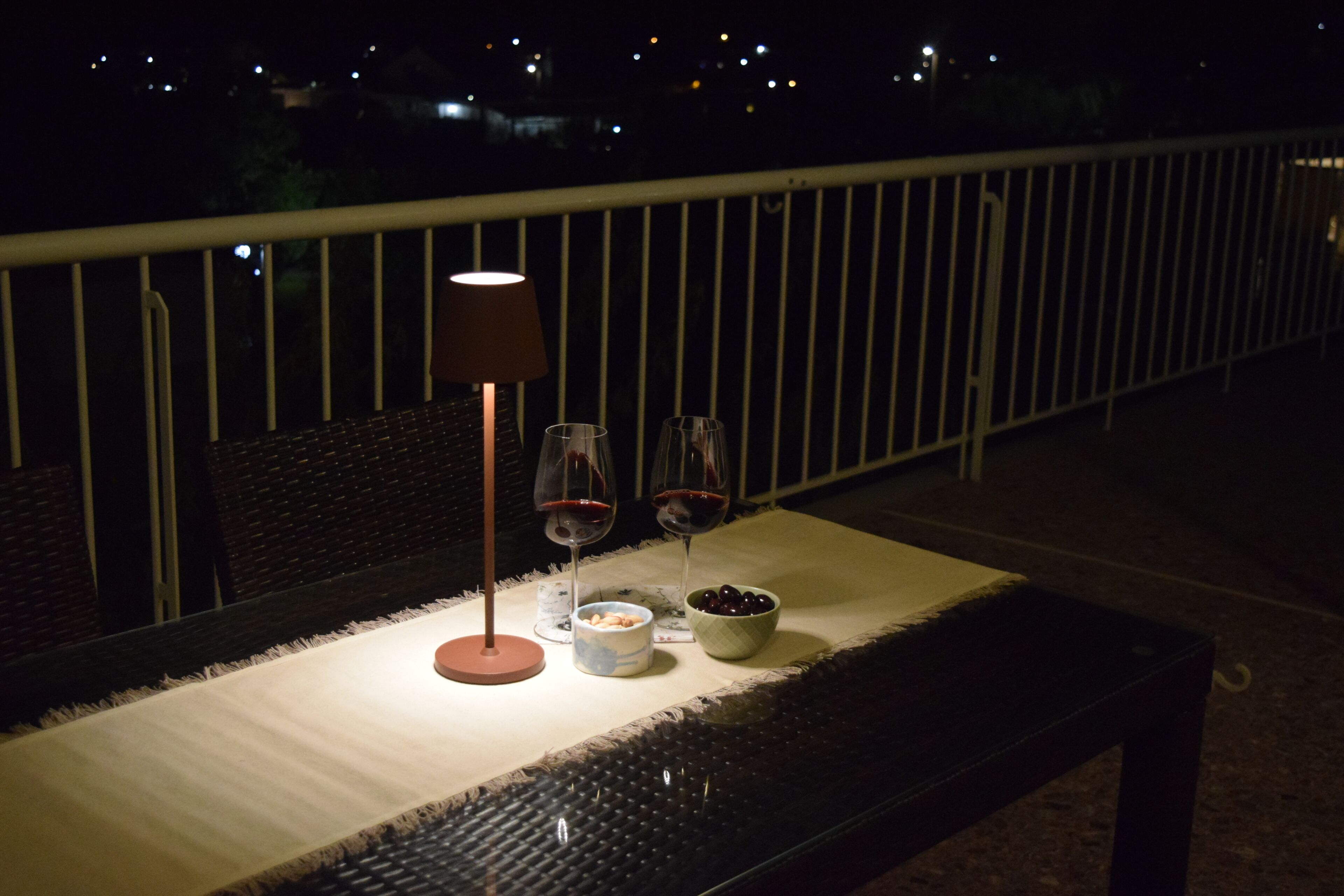 A balcony with a table set for two, featuring a lamp, two wine glasses, and a bowl of olives.