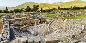 Un antiguo anfiteatro de piedra con asientos y un escenario central, rodeado de ruinas y colinas.