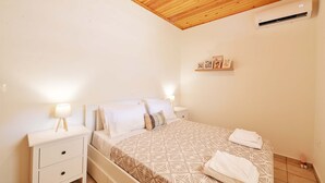 A bedroom with a wooden ceiling, a bed with a patterned bedspread, a bedside table with a lamp, and a wall-mounted air conditioner.