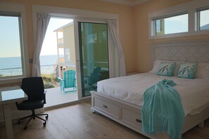 A bedroom with a bed, a desk, and a chair. There is a balcony with a view of the ocean.