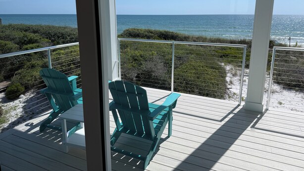Een balkon met twee groene Adirondack-stoelen en een witte tafel, met uitzicht op een strand, de oceaan en een heldere lucht.