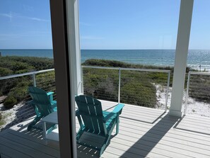 A balcony with two green Adirondack chairs and a white table, overlooking a beach with the ocean and a clear sky.