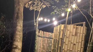 Un sendero de madera que atraviesa una valla de bambú, rodeado de árboles e iluminado con luces colgantes.
